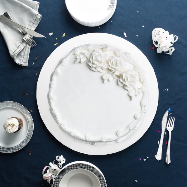 A white cake with white frosting on a Enjay white round cake drum on a table with silverware and cupcakes.