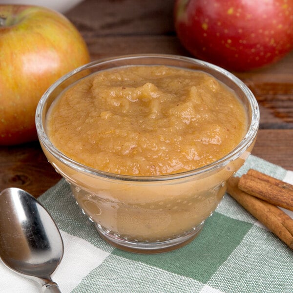 A glass bowl of Kime's applesauce with cinnamon sticks on a table.