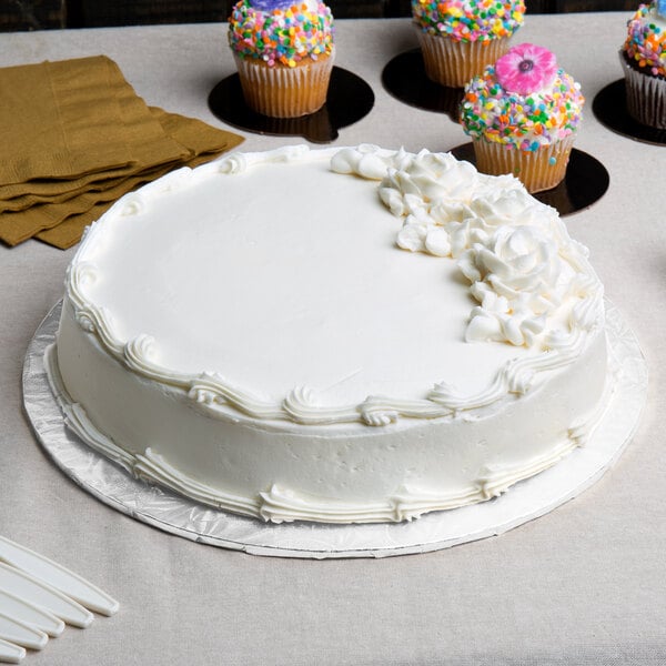 A white Enjay round cake drum under a frosted white cake on a table with cupcakes.