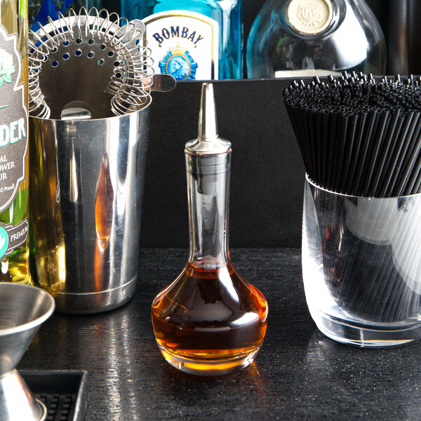 An American Metalcraft clear glass bitters bottle on a counter next to a glass of liquor.