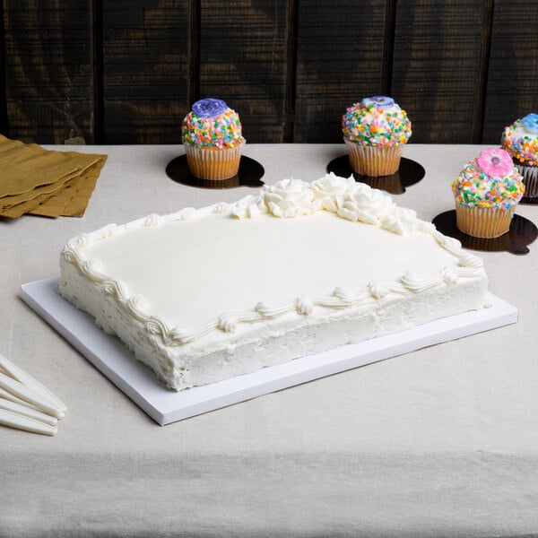 A white cake with frosting on a white Enjay quarter sheet cake board with cupcakes on a table.