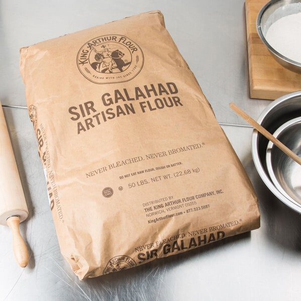 A brown bag of King Arthur Flour Sir Galahad Artisan Flour next to a bowl and rolling pin.