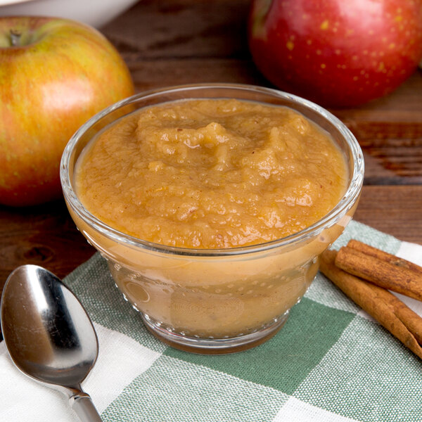 A glass bowl of Kime's cinnamon applesauce with a spoon on a napkin next to apples.