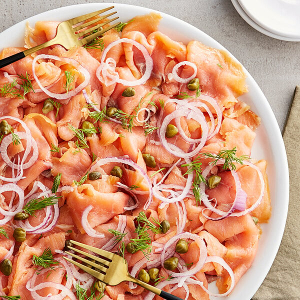 A plate of 3 lb. Atlantic smoked salmon with onions and pickles on a table in a deli with a fork.