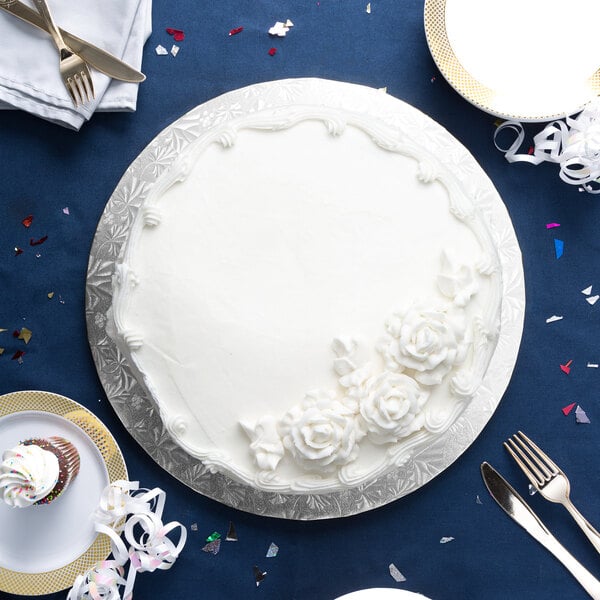 A white cake on a silver round cake board on a blue surface with silverware.