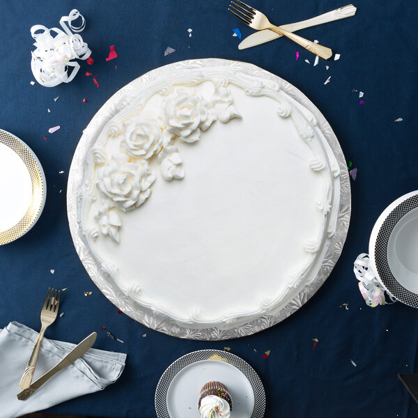 A white frosted cake on a silver round cake drum with a blue tablecloth.