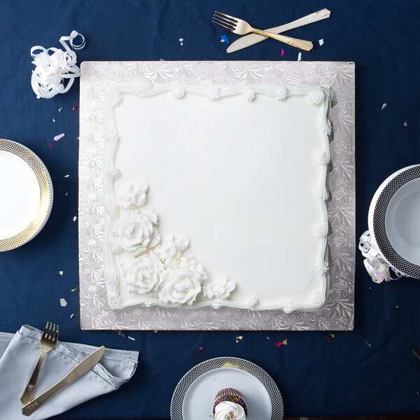 A white cake with white frosting on a silver square cake drum on a blue table.
