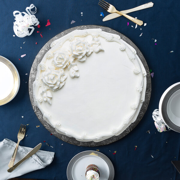 An Enjay black round cake drum under a white cake with white frosting and flowers on a blue table.