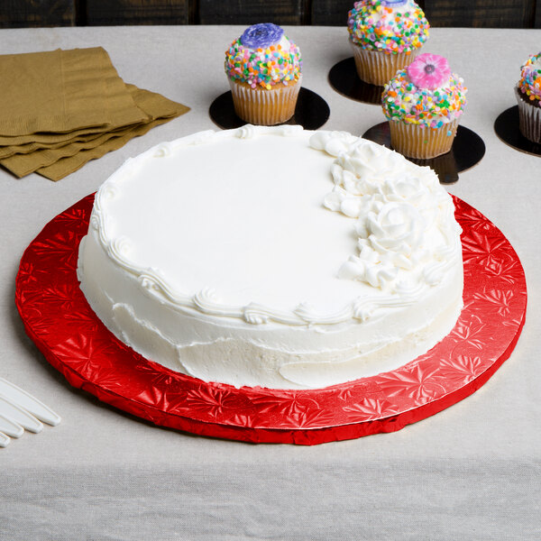A white frosted cake on a red round cake board on a table with cupcakes.