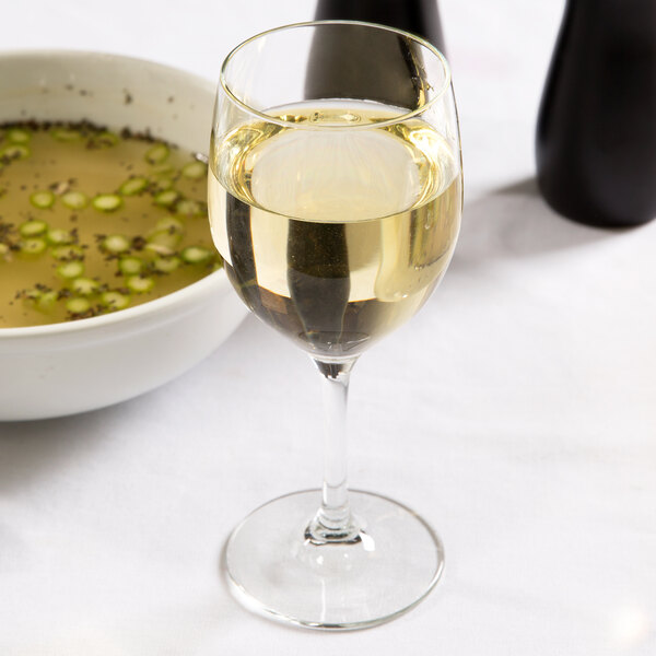 A Libbey Bristol Valley chalice wine glass filled with white wine on a table next to a bowl of soup.