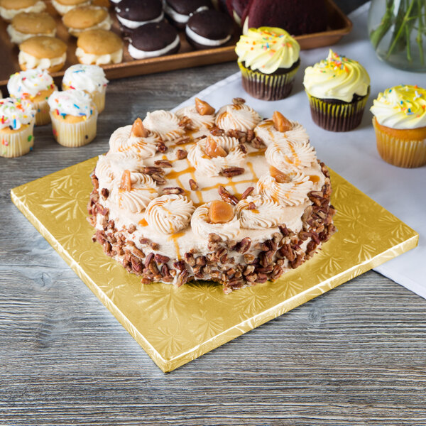 A cake with frosting and pecans on a gold Enjay cake drum on a table with cupcakes.