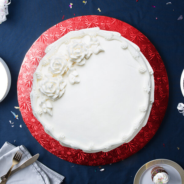 A white round cake with flowers on a red and white Enjay cake drum on a blue surface with a white plate, fork, and knife.