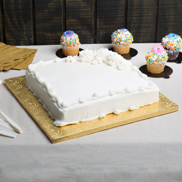 A white cake on a gold square cake drum with cupcakes on the table.