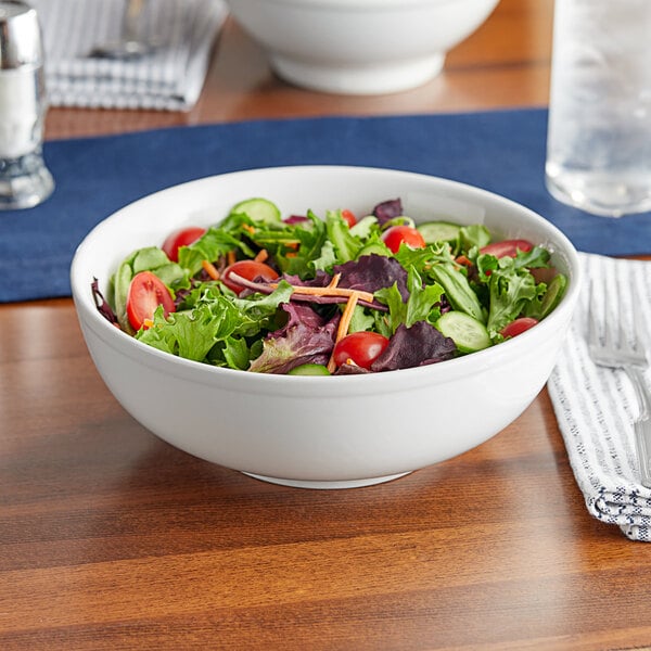 A close up of a white Acopa porcelain bowl filled with salad with tomatoes and cucumbers on a table.