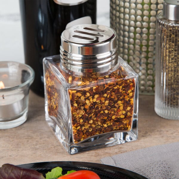 An American Metalcraft glass cheese shaker with a slotted stainless steel lid filled with red flakes on a table.