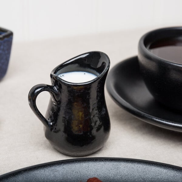 An American Metalcraft black porcelain bell creamer filled with white liquid on a table next to a cup of coffee.