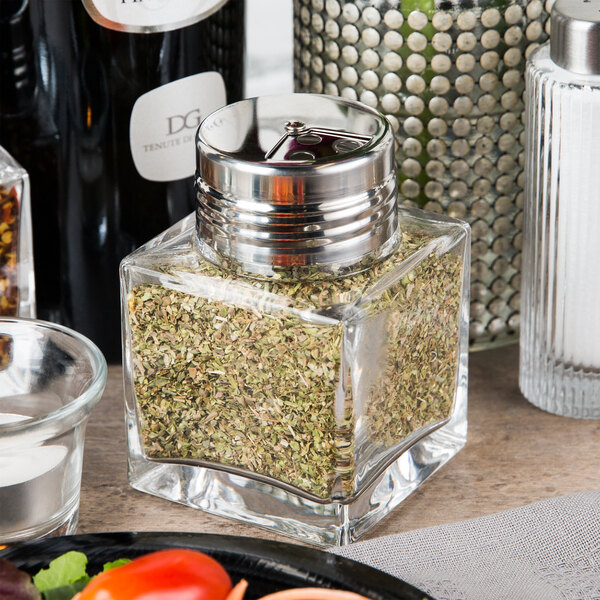An American Metalcraft square glass cheese/spice shaker with a stainless steel top filled with green herbs on a table.