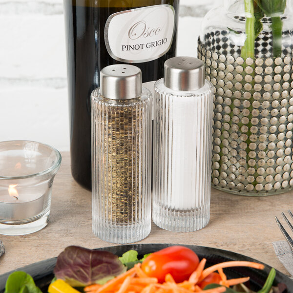 A table with a plate of vegetables and American Metalcraft ribbed round glass salt and pepper shakers next to a bottle of wine.