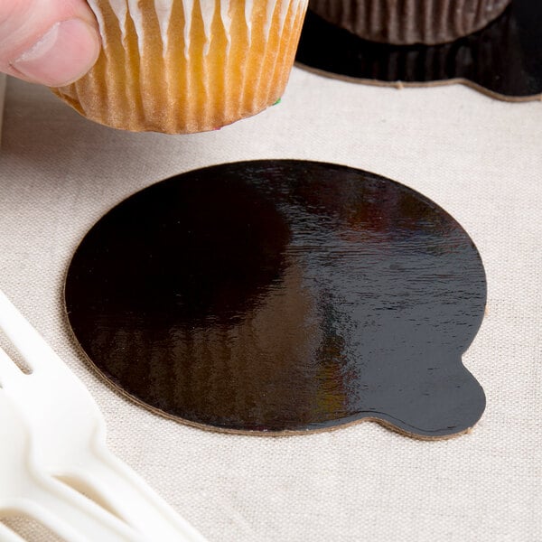 A person holding a cupcake with a black and gold Enjay round dessert board with a tab.