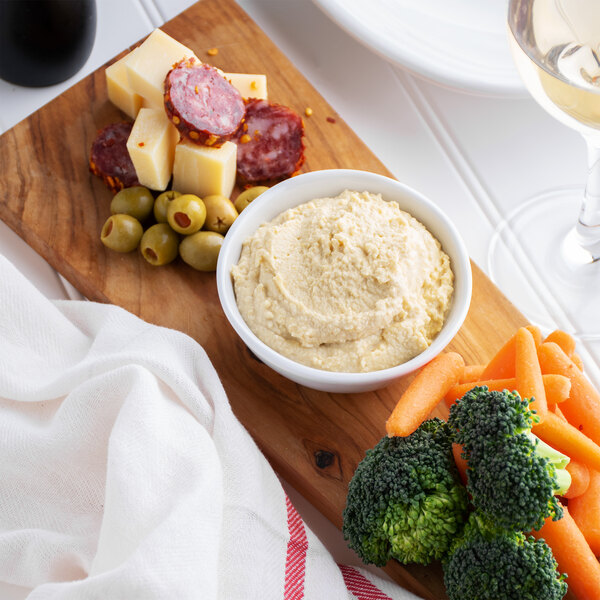 A bowl of Kronos classic hummus with olives, carrots, and broccoli on a table with a plate of food and a glass of wine.