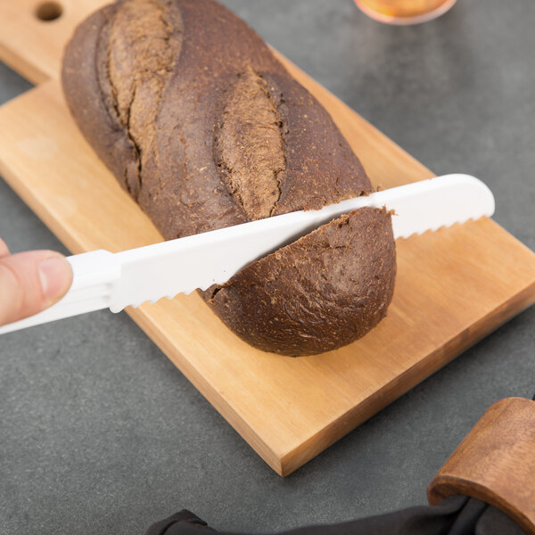 A person using a Fineline white plastic bread knife to cut bread on a cutting board.
