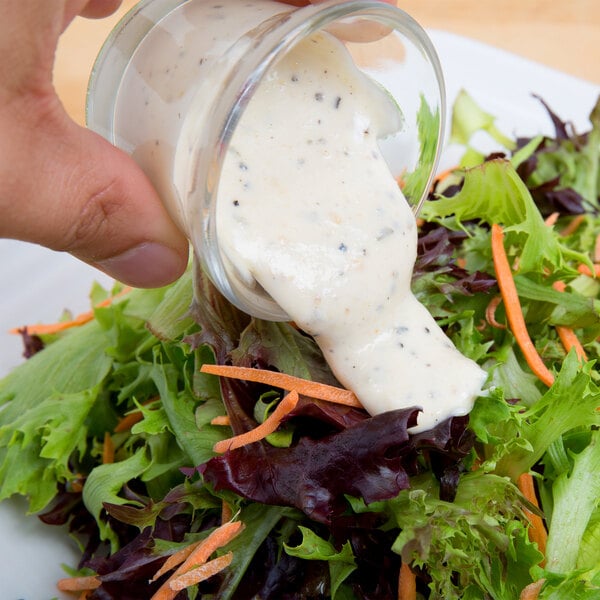 A hand pouring Ken's Foods Parmesan and Peppercorn dressing over a salad.
