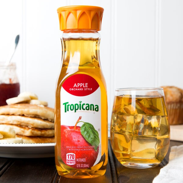A Tropicana bottle of apple juice next to a glass of apple juice with ice on a table.