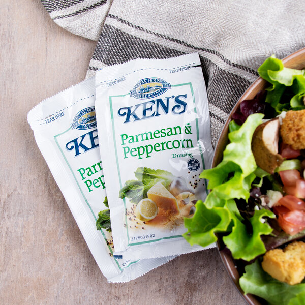 A bowl of salad with a Ken's Parmesan and Peppercorn dressing packet on a table with other condiments.
