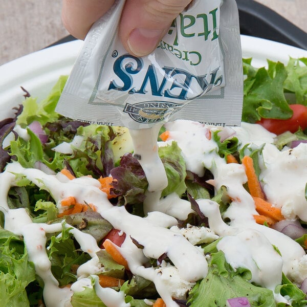 A hand pouring a Ken's Foods Creamy Italian dressing packet over a salad.