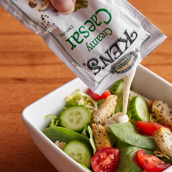 A person pouring a Ken's Foods Creamy Caesar dressing packet onto a bowl of salad.
