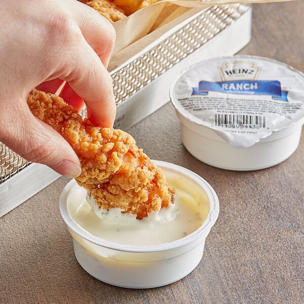 A hand dipping a fried chicken into a small container of Heinz Ranch Dressing.