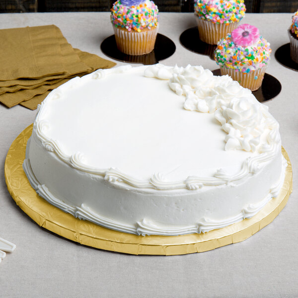 A white cake with frosting on a gold round cake drum with cupcakes on the table.