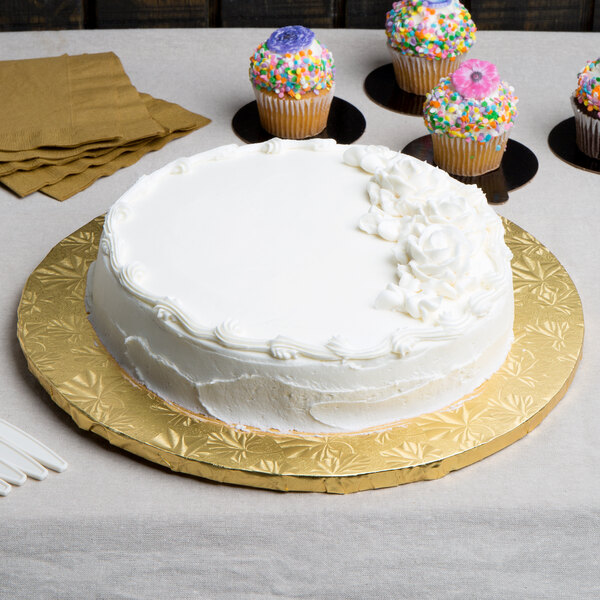 A white cake on a gold Enjay round cake drum with white frosting and cupcakes.