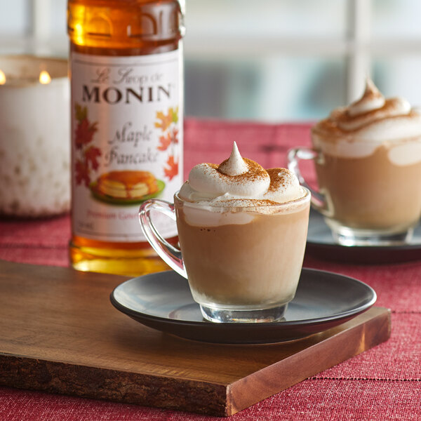 A glass cup of coffee with whipped cream and Monin syrup on a table with a bottle of Monin syrup.