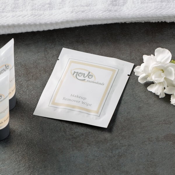 A white package of Novo Essentials Hotel and Motel Makeup Remover Wipes with a logo on it next to a white flower.