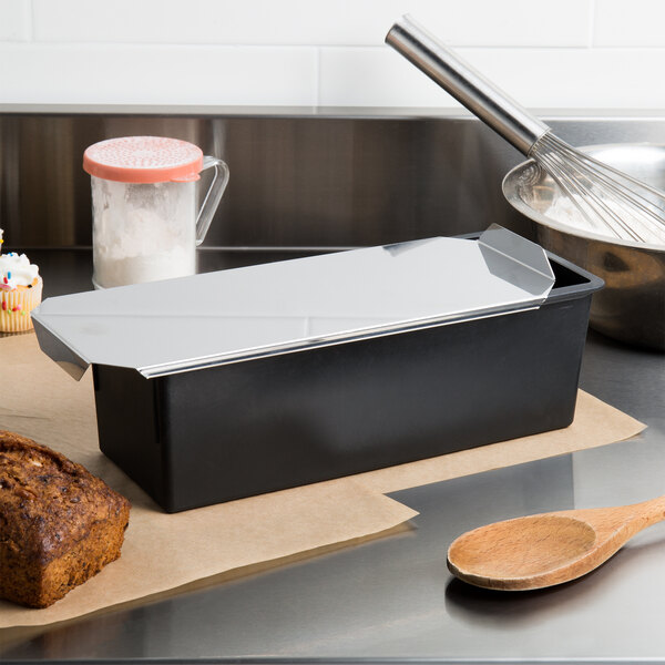 A loaf of bread in a black rectangular pan with a white lid on a counter.