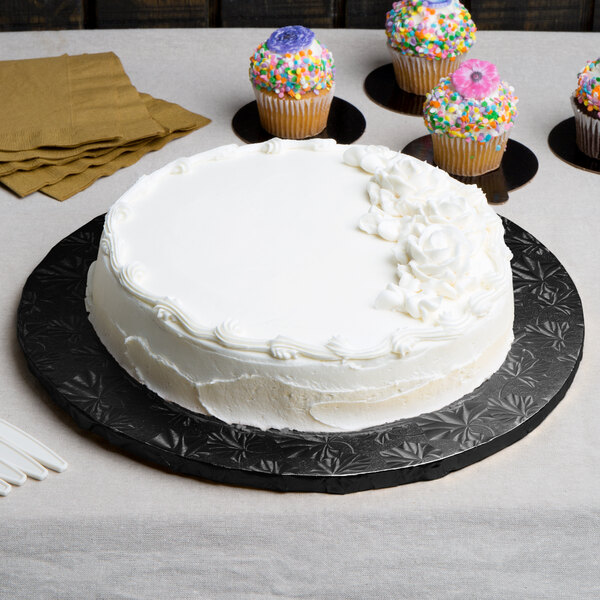 A white cake with frosting on a black Enjay round cake board with cupcakes on a table.