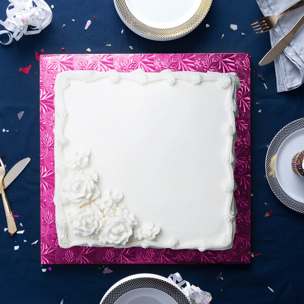 A white square cake on a pink cake drum with white flowers.