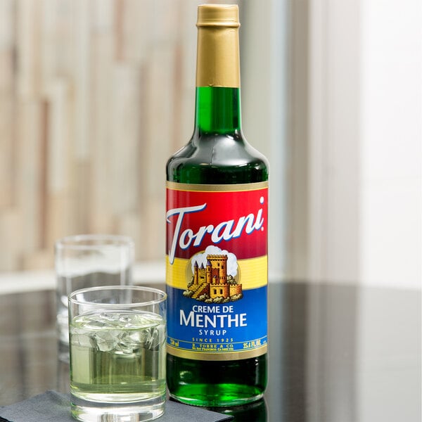 A Torani Creme de Menthe flavoring syrup bottle next to a glass of water with ice.