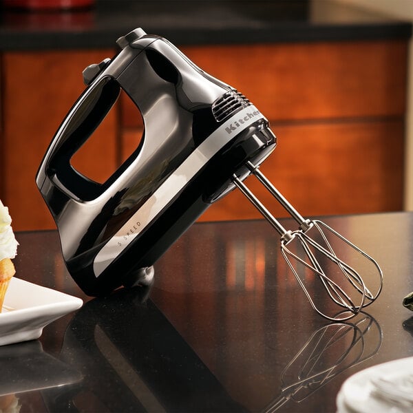 A black KitchenAid hand mixer with stainless steel beaters mixing batter on a white counter.