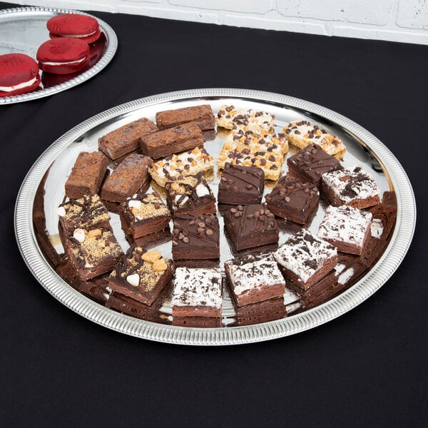 A stainless steel Vollrath round serving tray with a plate of brownies and cookies on a table.