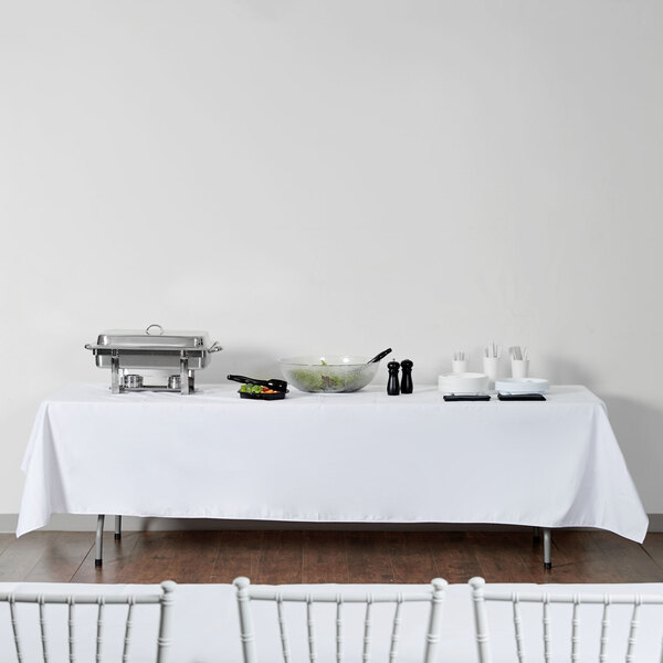 A rectangular table with a white Intedge tablecloth on it and silverware.