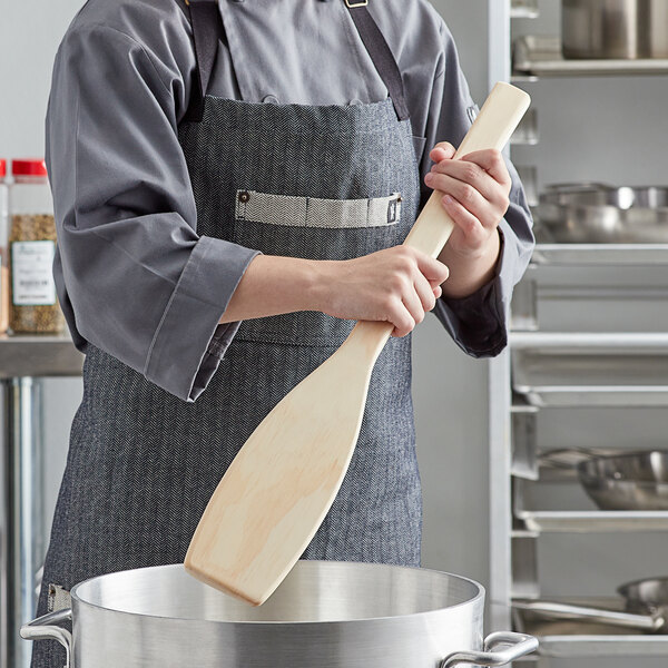 A person in a professional kitchen stirring a pot with an American Metalcraft wood paddle.