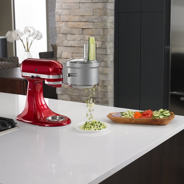 A red KitchenAid stand mixer with the KitchenAid food processor attachment being used to dice vegetables on a white counter.
