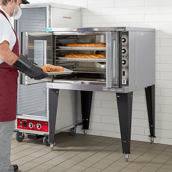 A man wearing a glove and mask holding a tray of bread in a Bakers Pride electric convection oven.