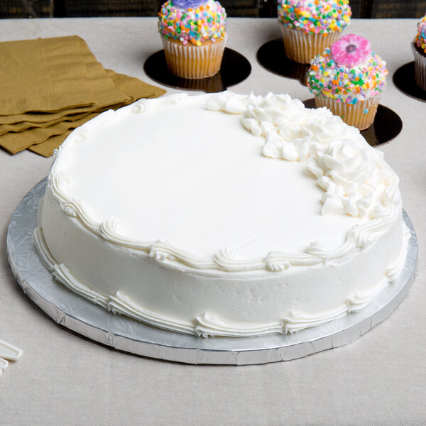 A white cake on an Enjay silver round cake drum on a table with cupcakes.