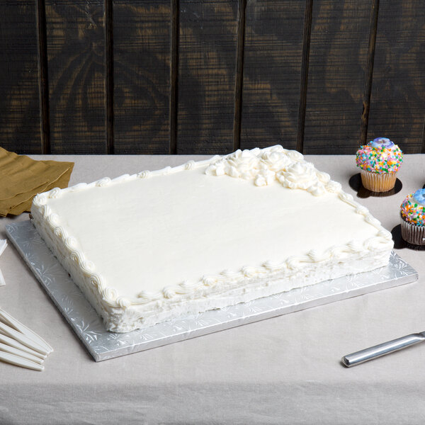 A white cake on an Enjay silver cake board on a table with cupcakes.