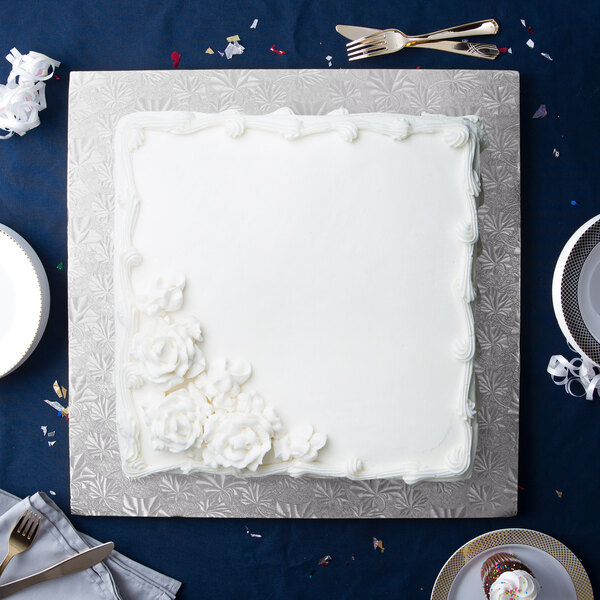 A white square cake on a silver Enjay cake drum with white frosting and flowers.