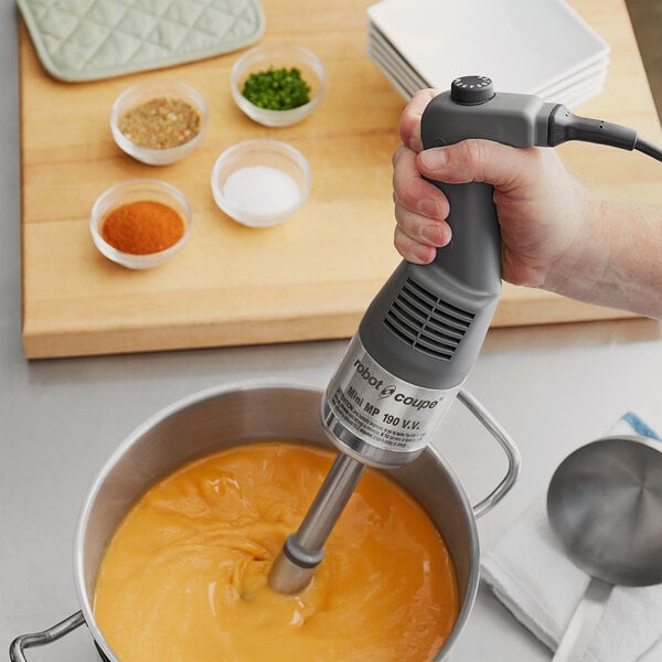 A hand using a Robot Coupe hand blender to mix a white sauce in a plastic bowl.