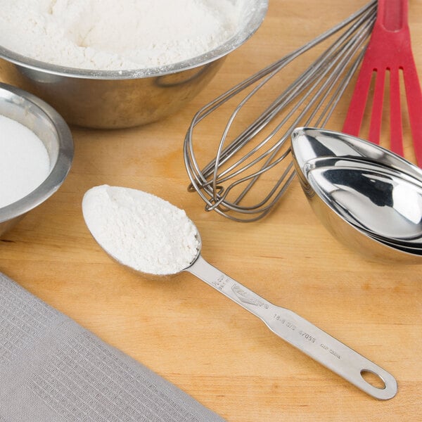 A Vollrath stainless steel measuring scoop with white powder in it next to a bowl of flour and a whisk.